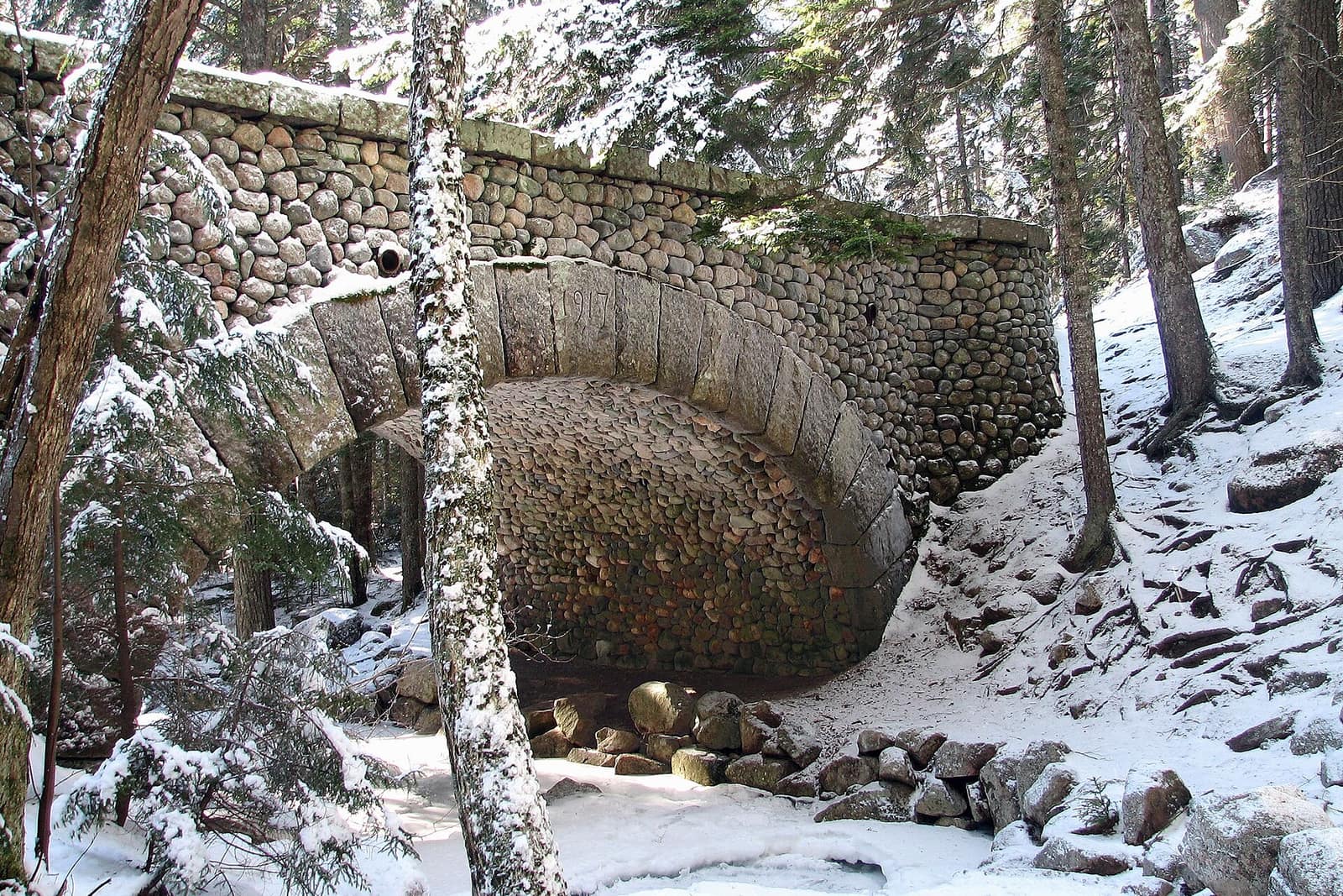 Snow-covered cobblestone carriage road bridge in Acadia National Park, Maine, surrounded by spruce trees in winter