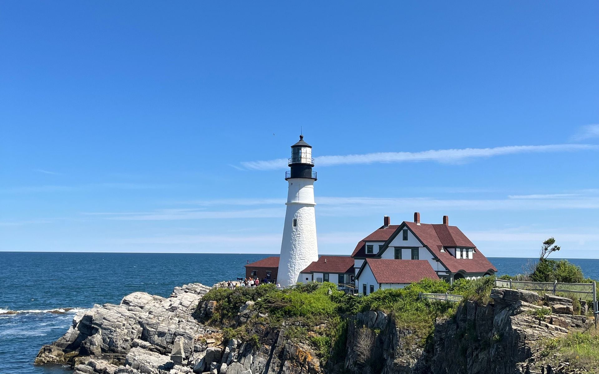 Two children exploring tide pools on a rocky Maine coastline with a lighthouse visible in the background on a clear summer day